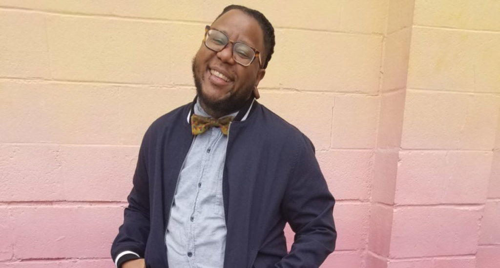 Amir Jones, a black person, wearing glasses, a bow tie and a cardigan poses and smiles at the camera