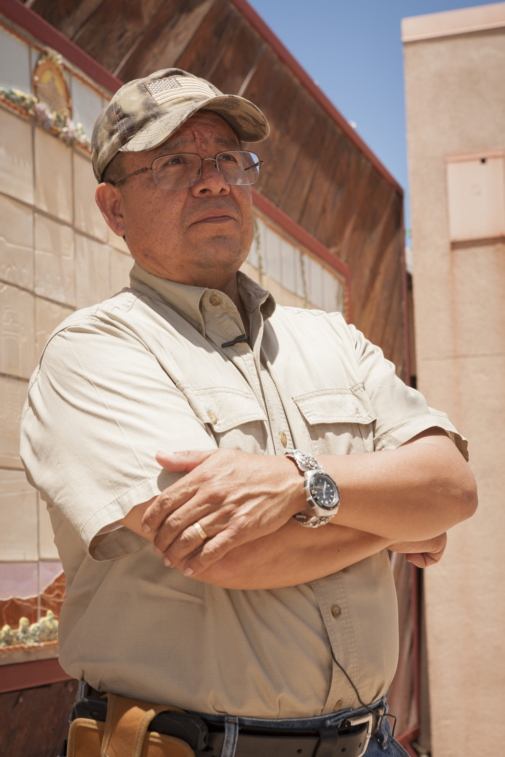 Emerson Toledo seen from the waist up, arms folded looking to the left, wearing a light brown khaki shirt, glasses and camouflage baseball cap.