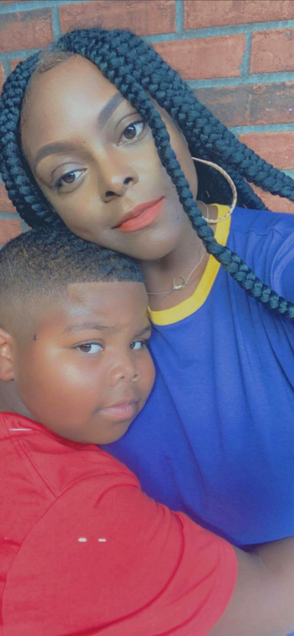 Selfie de Carnesia con su hijo frente a una pared de ladrillos. Lleva una camisa azul y tiene trenzas. Ella está apoyando la cabeza en su hijo, que tiene sus brazos alrededor de su cintura y su cabeza sobre su hombro. Lleva una camisa roja.