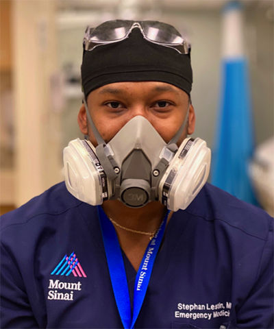 Dr. Lestin seen from the chest up facing the camera wearing dark blue scrubs, a respirator and goggles on top of his head.